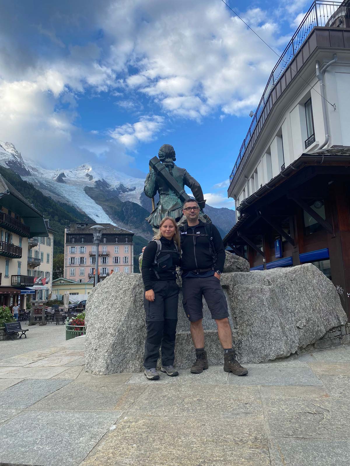 Two people standing in front of the Michel Paccard statue with Mont Blanc in the background
